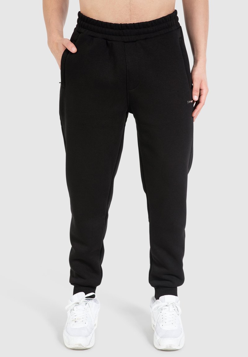 Schwarze Sweatpants mit einem elastischen Bund und Seitentaschen, aus einem weichen, strukturierten Stoff. Kombiniert mit weißen Sportschuhen.