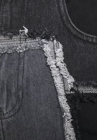 Tessuto in denim nero con bordi sfilacciati, cuciture grigie a contrasto e una superficie testurizzata con sezioni stratificate.