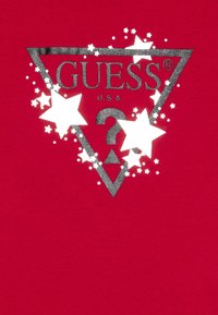 Czerwona bawełniana koszulka z trójkątnym, metalicznym srebrnym logo z napisem "GUESS" oraz rozproszonymi białymi gwiazdkami i dodatkowymi grafikami.