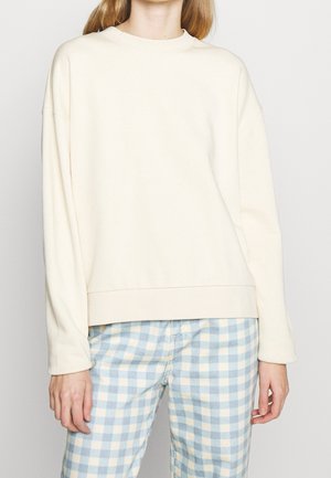 Person iført en ensfarvet off-white langærmet sweatshirt og lyseblå og hvide ternede bukser mod en hvid baggrund.