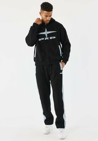 Sudadera con cremallera negra con logo "PXP", con paneles laterales grises. Combinada con pantalones de chándal negros y zapatillas blancas. Ropa atlética casual.
