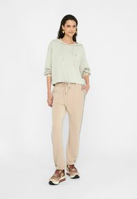 Sudadera de color claro con cordón y mangas acampanadas, combinada con joggers beige que tienen bolsillos laterales y ribetes. Zapatillas marrones con acentos texturizados.