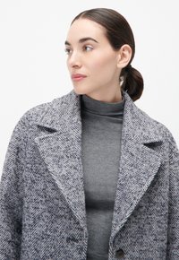 Grau gemusterter Herringbone-Mantel mit breiten Revers und Vorderknopfverschluss, getragen über einem grauen Rollkragenpullover, mit strukturiertem Stoff und lässiger Silhouette.
