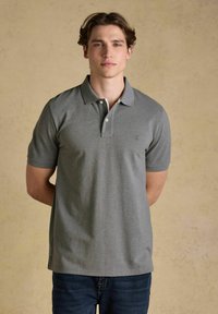 Noor mees seisab käed selja taga, seljas hall lühikeste varrukatega polo särk ja tumesinised teksad, taustaks beež sein.