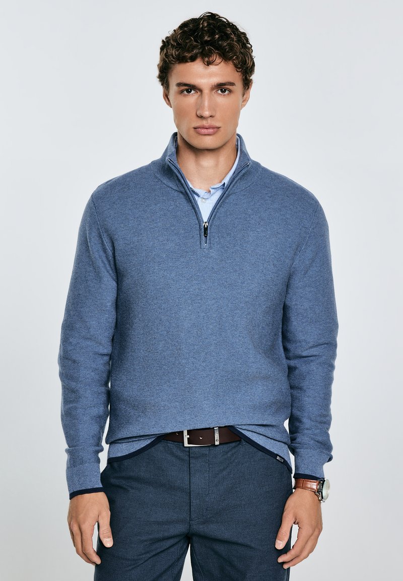 Un giovane uomo si erge con sicurezza, indossando un maglione blu con zip sopra una camicia con colletto, abbinato a pantaloni scuri e un orologio.