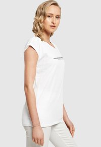 Hvid bomulds T-shirt med V-hals og opsnørede ærmer, med sort tekst på forsiden. Glat tekstur, lige snit, kombineret med lyse bukser.