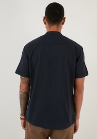 Cămașă scurtă, de culoare bleumarin închis, confecționată dintr-un material neted. Prezintă un guler clasic și o margine ușor curbată. Design simplu și versatil.
