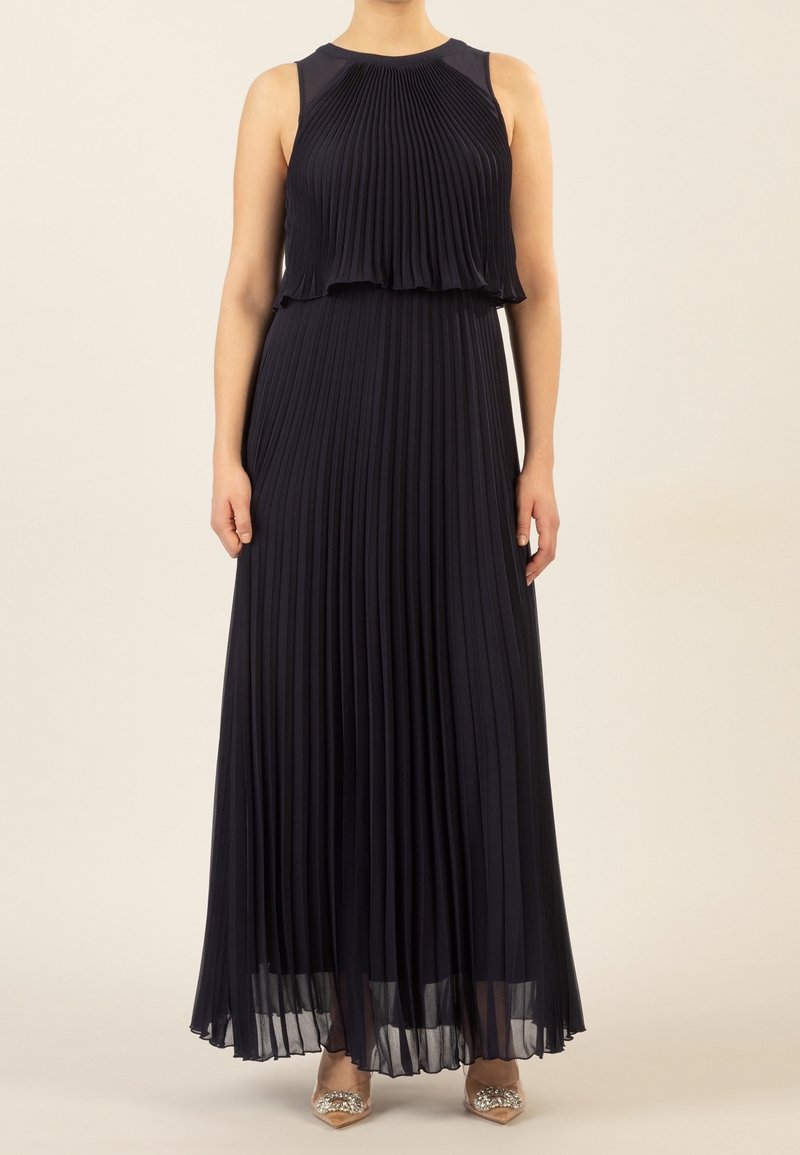 Femme portant une robe maxi plissée bleu marine sans manches avec un corsage à couches, debout devant un fond uni, visible du cou aux pieds.