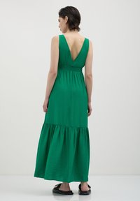 Frau in einem ärmellosen grünen Maxikleid mit tiefem V-Ausschnitt und gerafftem Bund, die vor einem schlichten hellen Hintergrund steht.