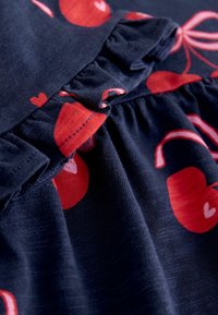 Temno modra tkanina z rdečimi vzorci sadja in src; ima nabrano spodnjico in naborke, ki poudarjajo mehko, teksturirano površino.