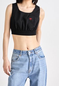 Haut court noir en tissu lisse avec un col rond dégagé et un ourlet élastique. Présente un petit détail de poche en forme de cœur rouge. Porté avec un jean bleu clair.