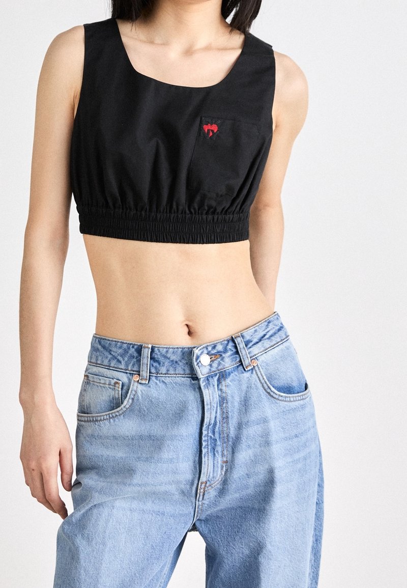 Haut court noir en tissu lisse avec un col rond dégagé et un ourlet élastique. Présente un petit détail de poche en forme de cœur rouge. Porté avec un jean bleu clair.