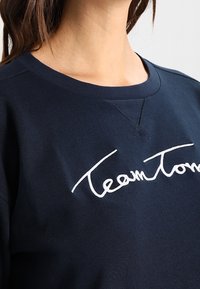 Námořnická modrá mikina s kulatým výstřihem, na hrudi s bílým vyšívaným textem "TeamTom". Měkká, strukturovaná látka.