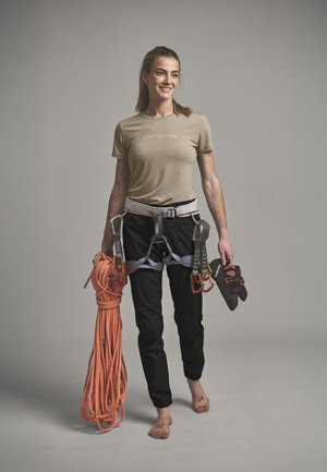 Frau trägt Klettergurt und Freizeitkleidung, hält in einer Hand ein aufgerolltes oranges Kletterseil und in der anderen Kletterschuhe, barfuß.