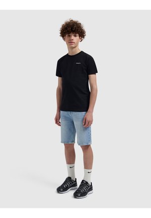 Tiener met krullend haar die een zwart T-shirt, lichtblauwe denim korte broek, witte Nike sokken en zwarte Nike sneakers draagt, staand op een witte achtergrond.