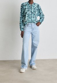 Vaaleansiniset leveälahkeiset farkut yhdistettyinä turkoosiin kuviolliseen puseroon, jossa on puffihihat ja pyöreä kaulus, pukeutuneena valkoisiin lenkkareihin.