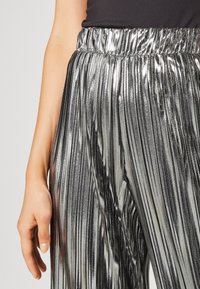 Metalické plisované kalhoty s elastickým pasem, které mají lesklou stříbrnou látku a hladkou strukturu. Detailní záběr na viditelné detaily látky.