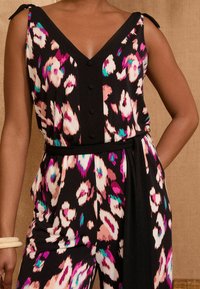 Črna brezrokavnica s potiskom abstraktnih cvetlic v roza, beli in zeleni barvi, gumbasto zapiranje spredaj, dekolte v obliki V, pas z vezalno vrvico in zanki na ramenih.