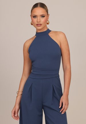 Mujer vestida con un mono sin mangas color azul marino con piernas anchas, pendientes de aro dorados y pulseras, sobre un fondo beige liso.