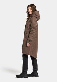 Braune wasserfeste Jacke mit Kapuze, Seitentaschen und Knopfdetails, kombiniert mit schwarzen Jeans und schwarzen Schnürstiefeln.