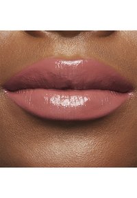 Nude-pink Lippenfarbe mit glänzendem Finish, kombiniert mit einer glatten Textur und voller Form, vor einem dunkleren Hautton.