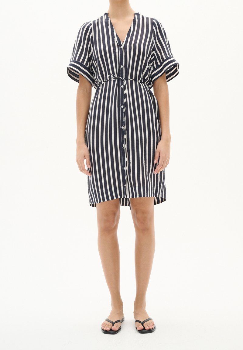 Femme portant une robe chemise rayée verticale bleu marine et blanc jusqu'aux genoux, avec des manches retroussées et une ceinture à la taille, associée à des sandales noires.
