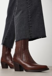 Botas de cuero marrón hasta el tobillo con punta afilada y tacón alto en bloque, con detalles de costura vertical. Combinadas con jeans de mezclilla negros deshilachados.