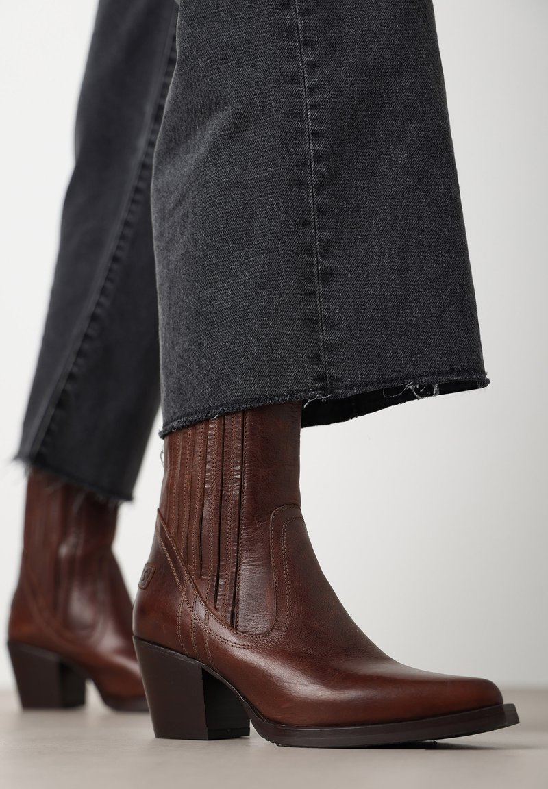 Botas de cuero marrón hasta el tobillo con punta afilada y tacón alto en bloque, con detalles de costura vertical. Combinadas con jeans de mezclilla negros deshilachados.