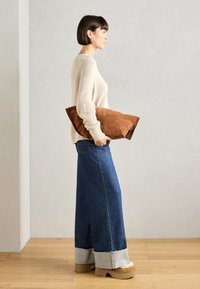 Overdimensjonert brun semsket skinnkrok holdt under armen; beige ribbestrikket genser og lang denimskjørt med lys denimkant; robuste brune plattform sko.