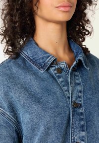 Jeans jakna v svetlo modri pranju s klasičnim ovratnikom, zapiranjem na gumbe in vidnimi šivi. Mehka tekstura, sproščen kroj.