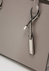Sac cabas gris clair texturé avec anses doubles et une étiquette métallique rectangulaire attachée par une lanière en cuir noir près du bord supérieur.