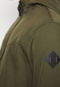 Oliivinvihreä hupullinen takki, jossa on sileä pinta, musta timantinmuotoinen laastari olkapäällä ja vetoketjusuljenta.