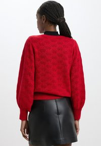 Maglione rosso lavorato a maglia con motivo a foglia testurizzato, maniche a sbuffo e orlo a coste. Il colletto nero contrasta con il colore vivace.