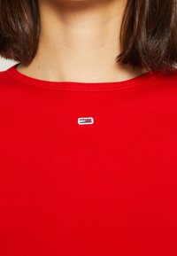 T-shirt rossa a costine con scollatura rotonda e un piccolo logo ricamato sul petto, con accenti blu, bianchi e neri.