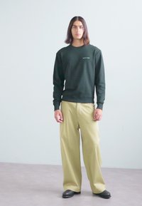 Dark green sweatshirt with a crew neck, paired with loose-fitting, light beige trousers. Model stands on a neutral background.
