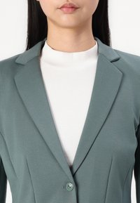 Sinine bleiser, millel on vormitud disain, sakilised kraed ja ühekohaline nööp üle valge ribakardiga turtlenecki. Sile tekstuur ja struktureeritud kuju.