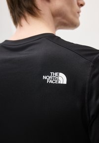 Férfi fekete, texturált rövid ujjú ingben, amelyen fehér "The North Face" logó látható a felső hátsó részen, kissé elfordulva a kamerától.