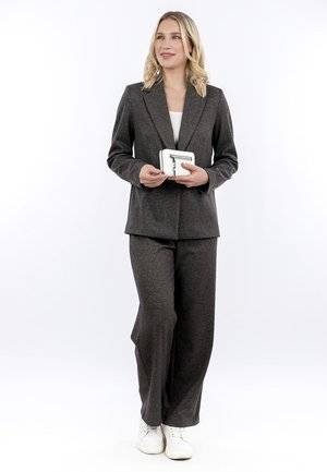 Vrouw staand in een donkergrijs pantalonpak met witte sneakers, een kleine witte portemonnee met ritssluiting vasthoudend, kijkt iets naar rechts.