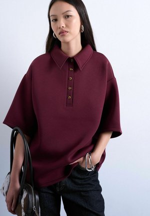 Ung kvinde med langt mørkt hår iført en vinrød kortærmet knapbluse, mørke jeans, hoops øreringe, armbånd og holder en sort håndtaske.