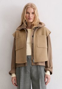 Beige cropped jack met grote kraag, ritssluiting aan de voorkant en twee borstzakken; gecombineerd met grijze geruite broek en een crèmekleurige top. Zachte textuur.