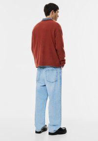 Rostfarbigem, gestricktem Pullover mit Rundhalsausschnitt, kombiniert mit hellblauen, oversized Jeans. Schwarz-platten Schuhe sichtbar.