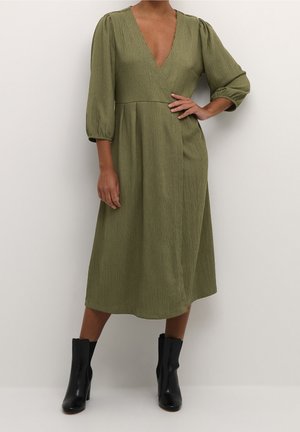 Woman wearing olive green midi dress with deep V-neck and three-quarter puff sleeves, paired with black ankle boots, standing against white wall.