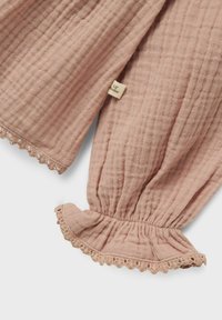 Svijetlo ružičasti muslin s dugim rukavima s volanima na manžetama i čipkastim detaljima. Uključuje malu oznaku brenda sa strane. Mekani, teksturirani materijal.