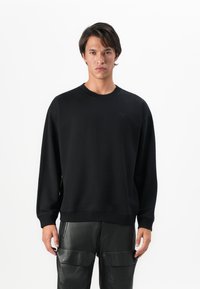 Un jeune homme portant un sweat-shirt noir uni et un pantalon en cuir noir, se tenant devant un fond blanc.