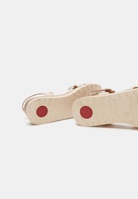 Sandales beiges avec une sangle texturée et une semelle épaisse. La semelle comporte un accent circulaire rouge et un motif de semelle pour une meilleure adhérence.