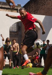 Piłkarz w czerwonej koszulce Puma i czarnych spodenkach uderza piłkę nożną. Wyraziste pomarańczowe korki i białe skarpetki podkreślają akcję.