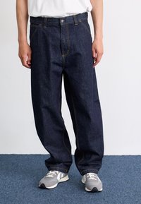 Dark blue denim jeans with a relaxed fit, featuring yellow stitching, front pockets, and a metal button closure. Worn with gray sneakers.