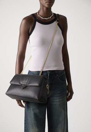 Black leather handbag with a structured design, flap closure, gold-tone chain strap, and geometric hardware detail. Worn over a light tank top.