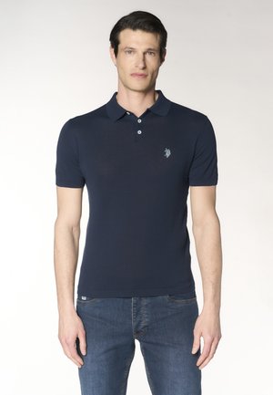 Uomo che indossa una polo blu navy aderente a maniche corte con un piccolo logo ricamato sul petto e jeans blu.