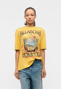 Γυναίκα με πλεξούδες που φοράει κίτρινο φαρδύ μπλουζάκι με πολύχρωμο σχέδιο «Billabong 1973 World Tour» και άνετο μπλε τζιν.